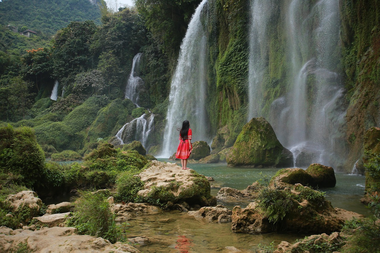 Une visiteuse en robe rouge au pied d'une chute latérale de Ban Gioc, donnant l'échelle du rideau d'eau