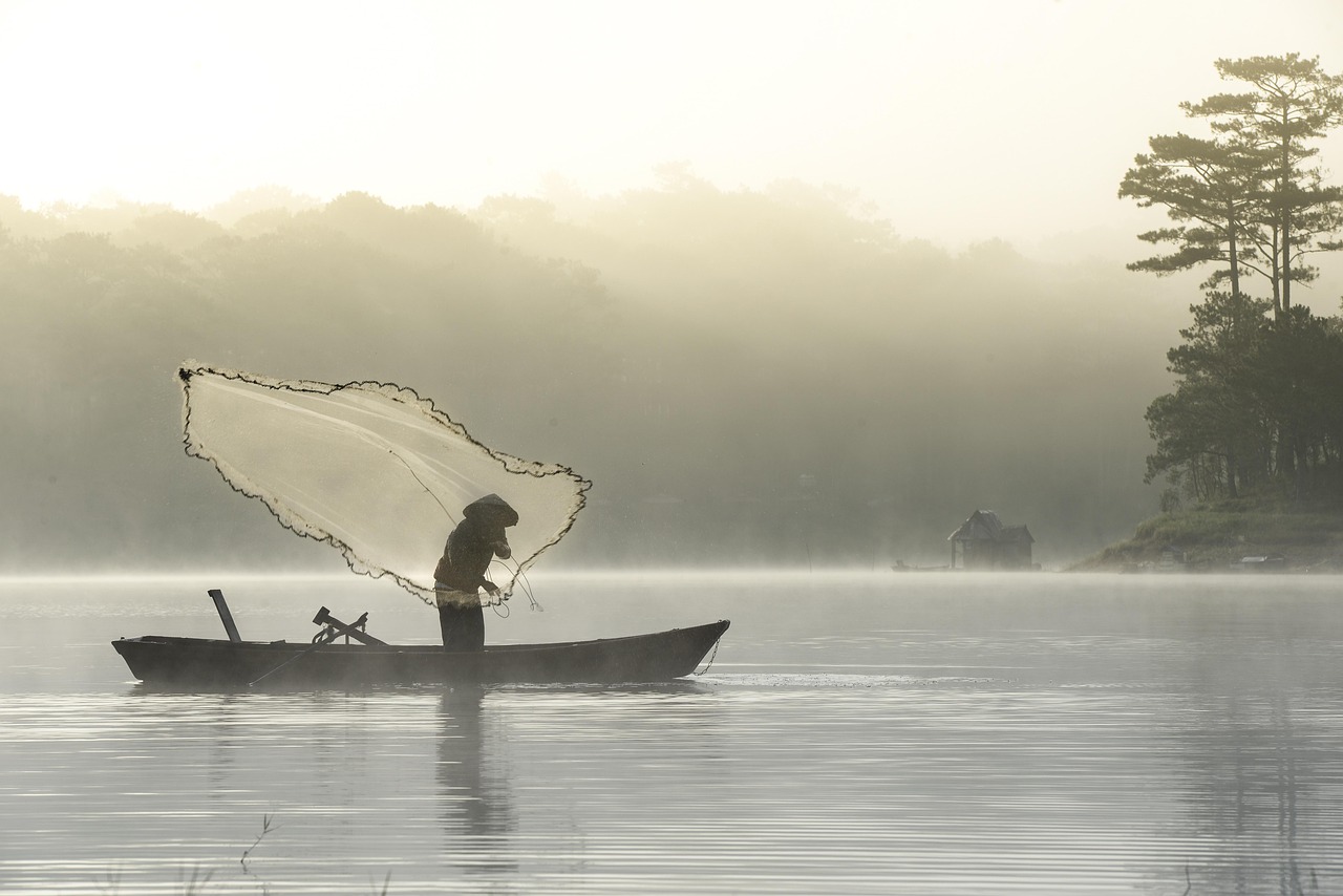 A fisherman casting his net from a boat on a lake shrouded in morning mist near Đà Lạt
