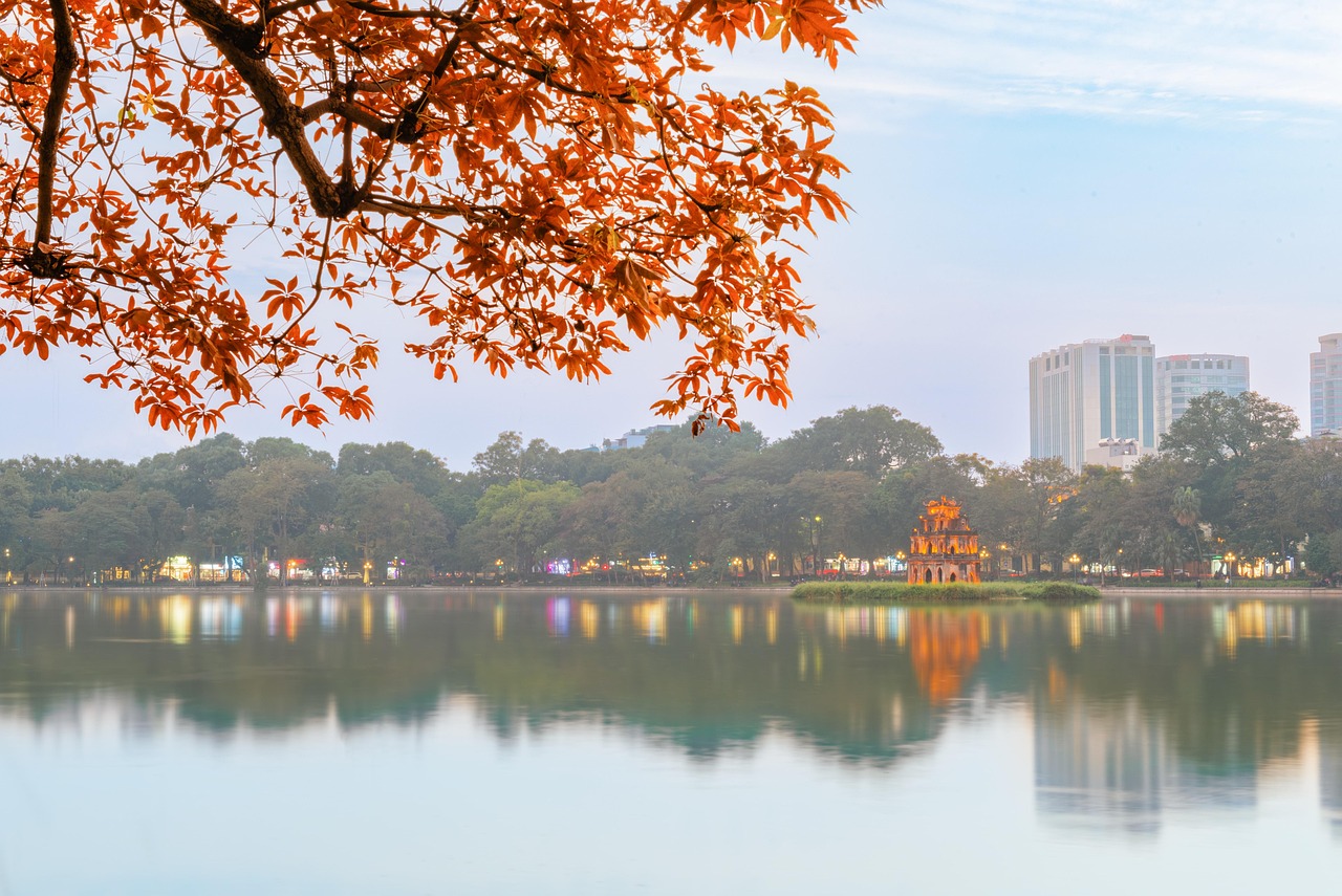 Hoàn Kiếm Lake at dusk in autumn: the Turtle Tower lit up on its islet and city lights reflected in the water, beneath a branch of orange leaves