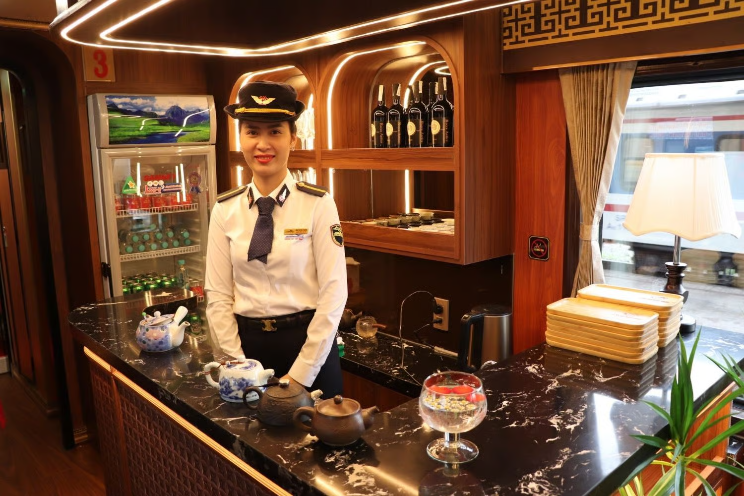 Hôtesse en uniforme blanc derrière le bar à thé en marbre noir d'une voiture de service du train Huế – Phong Nha, avec théières et vitrine éclairée