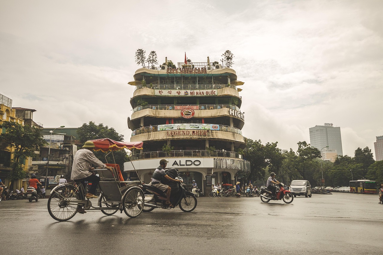 Cyclo and motorbikes crossing a Hanoi Old Quarter intersection under overcast skies after a rain shower